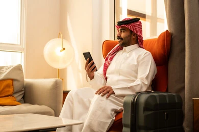 إجراء بث مباشر مع الأهل والأصدقاء، استخدام الهاتف المحمول، دمج التقنيات الحديثة فى الحياة اليومية، تصفح مواقع التواصل الاجتماعي ، رجل عربي خليجي سعودي الثوب الابيض و الشماغ يجلس في الفندق بجانب حقيبة السفر يمسك الجوال بيده ، التقاط صور السيلفي التذكارية