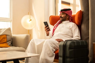 استخدام الهاتف المحمول، دمج التقنيات الحديثة فى الحياة اليومية، تصفح مواقع التواصل الاجتماعي ، رجل عربي خليجي سعودي الثوب الابيض و الشماغ يجلس في الفندق بجانب حقيبة السفر يمسك الجوال بيده ، إجراء بث مباشر مع الأهل والأصدقاء، التقاط صور السيلفي التذكارية