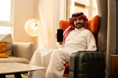 Expressions of happiness and joy, using modern devices and technologies, a Saudi Arabian Gulf man in a white thobe and ghutrah sitting in a hotel next to a suitcase holding a mobile phone in his hand, displaying a blank black screen with gestures of joy.