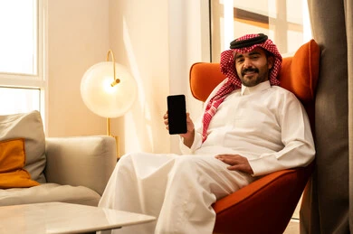 عرض شاشة سوداء فارغة بإيماءات السرور، تعابير السعادة والفرح، استخدام الأجهزة والتقنيات الحديثة، رجل عربي خليجي سعودي الثوب الابيض و الشماغ يجلس في الفندق يمسك الجوال بيده