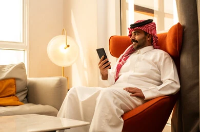 دمج التقنيات الحديثة فى الحياة اليومية، تصفح مواقع التواصل الاجتماعي ، رجل عربي خليجي سعودي الثوب الابيض و الشماغ يجلس في الفندق يمسك الجوال بيده ، استخدام الهاتف المحمول ، إجراء بث مباشر مع الأهل والأصدقاء، التقاط صور السيلفي التذكارية