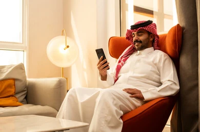 Browsing social media, an Arab Gulf Saudi man in a white thobe and ghutrah sits in a hotel holding a mobile phone in his hand, integrating modern technologies into daily life, using a mobile phone, conducting a live broadcast with family and friends, taking commemorative selfies.
