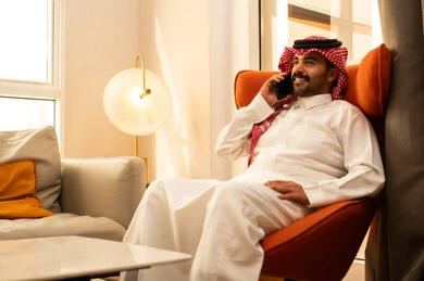 Exchanging conversations and good news, making a voice call via mobile, an Arab Gulf Saudi man wearing a white thobe and a ghutrah sitting in a hotel using a mobile phone, gestures of happiness while communicating with friends and relatives.