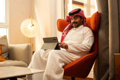 Browsing social media, watching enjoyable movies and series, using modern technology and techniques, an Arab Gulf Saudi man wearing a white thobe and a ghutrah sits in a hotel using a tablet.