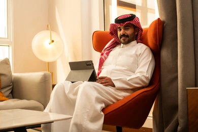 متابعة الأفلام والمسلسلات الممتعة، تصفح مواقع التواصل الاجتماعي, استخدام التكنولوجيا والتقنيات الحديثة، رجل عربي خليجي سعودي يرتدي الثوب الابيض و الشماغ يجلس في الفندق يستخدم التابلت