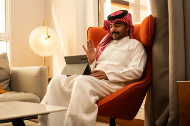 Raising the hand with gestures of greeting and peace, making a video call, browsing social media, using modern technology and techniques, an Arab Gulf Saudi man wearing a white thobe and a ghutrah sitting in a hotel using a tablet.