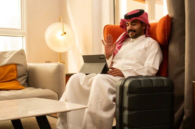 اجراء مكالمة فيديو، رفع كف اليد بيتابير التحية و السلام، تصفح مواقع التواصل الاجتماعي, استخدام التكنولوجيا والتقنيات الحديثة، رجل عربي خليجي سعودي يرتدي الثوب الابيض و الشماغ يجلس في الفندق بجانب حقيبة السفر يستخدم التابلت