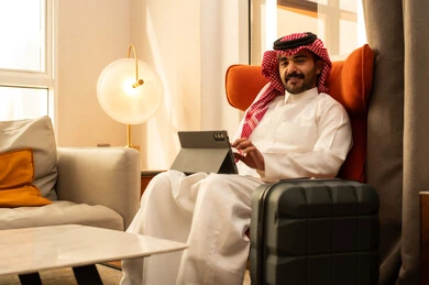 Watching enjoyable movies and series, browsing social media, using modern technology and techniques, an Arab Gulf Saudi man wearing traditional attire and a ghutrah sits in a hotel next to a travel bag using a tablet.