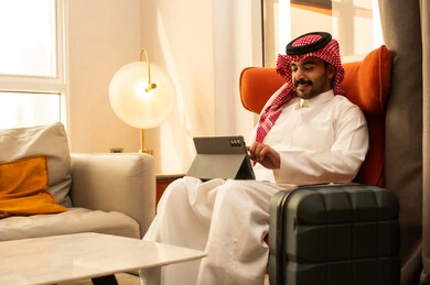 Browsing social media, using modern technology and techniques, an Arab Gulf Saudi man wearing traditional attire and a ghutrah sits in a hotel next to a travel bag using a tablet, watching enjoyable movies and series.
