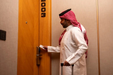 Using the smart card to open the room door, enjoying the holiday vacation, a modern and advanced lock in a luxury hotel, an Arab Gulf Saudi man wearing a white thobe and a shemagh holding a suitcase in his hands is opening the door of a hotel room, tourism in the Kingdom of Saudi Arabia.