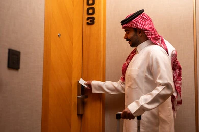 Enjoying a tourist vacation, a modern and advanced lock in a luxury hotel, an Arab Gulf Saudi man wearing a white thobe and a shemagh holding a suitcase in his hands is opening the door to a hotel room, tourism in the Kingdom of Saudi Arabia, opening the door with an electronic card.