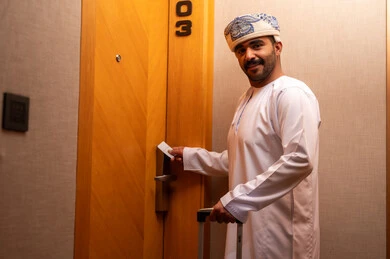 Looking at the camera with expressions of joy, a modern and advanced lock in a luxury hotel, an Arab Gulf Omani man wearing a dishdasha and turban is opening the door to a hotel room, tourism in the Kingdom of Saudi Arabia, opening the door with an electronic card, enjoying tourist holidays and the summer vacation.