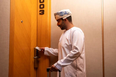 A Gulf Arab Omani man wearing a dishdasha and a turban is opening the door to a hotel room, tourism in the Kingdom of Saudi Arabia, a modern and advanced lock in a luxury hotel, opening the door with an electronic card, enjoying tourist holidays and the summer vacation.