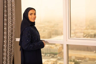 Confidence and self-esteem, attention to elegance and appearance, the concept of femininity and softness, a smiling, veiled Saudi Gulf Arab woman wearing an abaya stands by the window looking at the camera with gestures of joy and happiness, with the background of towers and skyscrapers in Riyadh.