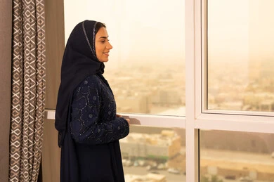 الثقة والاعتزاز بالنفس، الاهتمام بالأناقة والمظهر الخارجي، مفهوم الأنوثة والنعومة، امرأة عربية خليجية سعودية مبتسمة محجبة ترتدي عباءة تقف بجانب النافذة  بإيماءات السرور والسعادة، خلفية الابراج وناطحات السحاب في الرياض