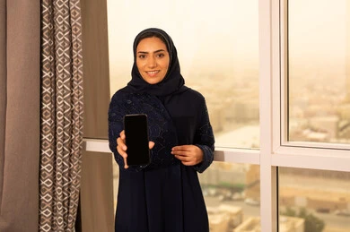 Looking at the camera with gestures of happiness and joy, showcasing mobile applications, an Arab Gulf Saudi woman wearing a black abaya stands by the window holding a mobile phone in her hand, using a modern and advanced technological device.
