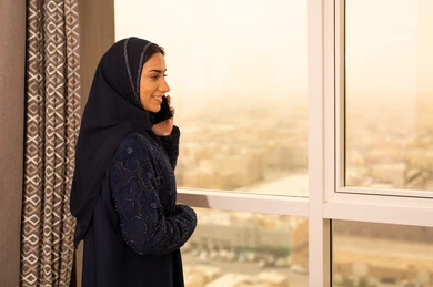 Making a voice call via mobile phone, conversations and chats with family and relatives, using modern technologies and advanced technology, a side view of a Gulf Arab Saudi woman wearing an abaya and hijab standing by the window holding a mobile phone in her hand.
