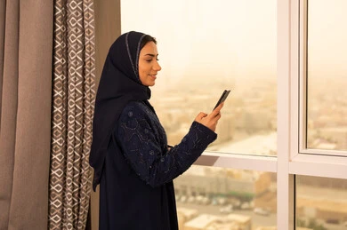 الدردشة مع الأصدقاء والأقارب عبر الجوال، تصفح مواقع التواصل الاجتماعي، استخدام جهاز تقني حديث ومتطور، صورة من الجانب لامراة عربيه خليجية سعودية ترتدي العباءة و الحجاب تقف بجانب النافذة تمسك بيدها الهاتف المحمول
