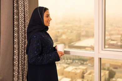 Gestures of enjoyment and relaxation, enjoying hot beverages, spending free time sipping a favorite drink, consuming warm winter herbs, a side view of a Saudi Gulf Arab woman wearing an abaya and hijab standing by the window.