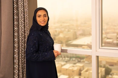 Enjoying hot drinks, gestures of pleasure and relaxation, spending free time sipping a favorite beverage, consuming warm winter herbs, an Arab Gulf Saudi woman wearing an abaya and hijab standing by the window. Enjoying hot drinks, gestures of pleasure and relaxation, spending free time sipping a favorite beverage, consuming warm winter herbs, an Arab Gulf Saudi woman wearing an abaya and hijab standing by the window.