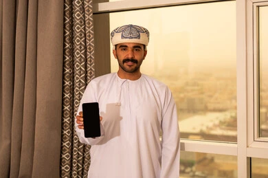 A blank black screen is displayed on the mobile phone, looking at the camera with gestures of happiness, using modern and advanced technology, an Arab Gulf Omani man wearing a dishdasha and kumma stands by the window holding the mobile phone in his hand.