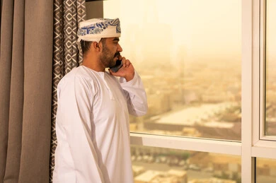 حوارات ودردشات مع الاهل والاقارب، اجراء مكالمة صوتية عبر الهاتف المحمول، استخدام التقنيات الحديثة والتكنولوجيا المتطورة،رجل عربي خليجي عماني يرتدي الدشداشة و الكمة يقف بجانب النافذة يحمل بيده الجوال