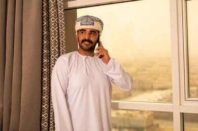 Making a voice call via mobile phone, conversations and chats with family and relatives, using modern technologies and advanced technology, an Arab Gulf Omani man wearing a dishdasha and a kumma stands by the window holding a mobile phone in his hand.