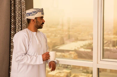 Relaxing and having warm drinks, trying to get a little rest, enjoying a cup of hot tea, an Arab Gulf Omani man wearing a dishdasha and a kumma stands by the window holding a cup of his favorite drink.