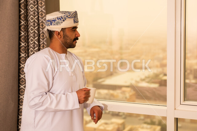 محاولة الحصول على القليل من الراحة، التلذذ بكوب من الشاي الساخن، الاسترخاء وتناول مشروبات دافئة، رجل عربي خليجي عماني يرتدي  الدشداشة و الكمة يقف بجانب النافذة يمسك بيده فنجان من مشروبه المفضل