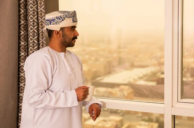 محاولة الحصول على القليل من الراحة، التلذذ بكوب من الشاي الساخن، الاسترخاء وتناول مشروبات دافئة، رجل عربي خليجي عماني يرتدي  الدشداشة و الكمة يقف بجانب النافذة يمسك بيده فنجان من مشروبه المفضل