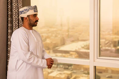 التلذذ بكوب من الشاي الساخن، محاولة الحصول على القليل من الراحة، الاسترخاء وتناول مشروبات دافئة، رجل عربي خليجي عماني يرتدي  الدشداشة و الكمة يقف بجانب النافذة يمسك بيده فنجان من مشروبه المفضل