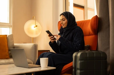 Gestures of integration and focus, benefiting from modern technological devices during leisure time, a Gulf Arab Saudi woman wearing an abaya and hijab sitting inside a hotel next to a suitcase in front of a laptop, using a mobile phone for chatting and communicating with family and friends.