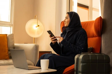 النظرالى شيء ما، الاستفادة من الأجهزة التكنولوجية الحديثة في أوقات الفراغ، امرأة عربية خليجية سعودية ترتدي العباءة والحجاب تجلس داخل الفندق بجانب حقيبة السفر امام الحاسوب المحمول، استخدام الجوال في الدردشة والتواصل مع الأهل و الأصدقاء