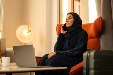 Making a voice call via mobile, laughing and chatting with friends, communicating and chatting with family and relatives, an Arab Gulf Saudi woman wearing a hijab and abaya sitting in a hotel next to a suitcase, sitting in front of a laptop holding a mobile phone in her hands.