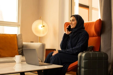 الضحك والتسامر مع الاصدقاء، اجراء مكالمة صوتية عبر الجوال، التواصل والدردشة مع الاهل والاقارب، امراة عربيه خليجية سعودية محجبة ترتدي العباءة و الحجاب تجلس فى الفندق بجانب حقيبة السفر تجلس امام الحاسب المحمول تحمل بيديها الهاتف المحمول