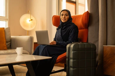 إنجاز المهام في عن بعد، ايماءات السعادة و السرور، مهن ووظائف نسائية، دمج التكنولوجيا بالعمل، ادارة الأعمال عن بعد، إمرأة عربية خليجية سعودية ترتدي العباءة والحجاب تجلس داخل الفندق بجانب حقيبة السفرتستخدم الحاسوب المحمول