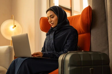 Gestures of focus and engagement, women's professions and jobs, integrating technology into work, remote business management, an Arab Gulf Saudi woman wearing an abaya and hijab sitting inside a hotel next to a travel bag using a laptop, accomplishing tasks remotely.