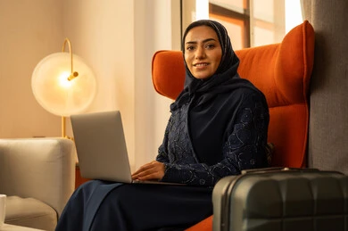 ايماءات السعادة و السرور، إنجاز المهام في عن بعد، مهن ووظائف نسائية، دمج التكنولوجيا بالعمل، ادارة الأعمال عن بعد، إمرأة عربية خليجية سعودية ترتدي العباءة والحجاب تجلس داخل الفندق بجانب حقيبة السفرتستخدم الحاسوب المحمول