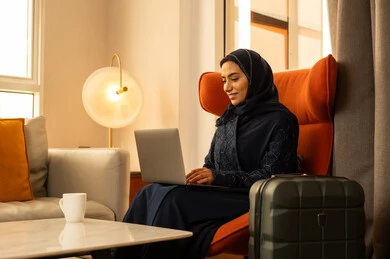 مهن ووظائف نسائية، ايماءات التركيز والاندماج، دمج التكنولوجيا بالعمل، ادارة الأعمال عن بعد، إمرأة عربية خليجية سعودية ترتدي العباءة والحجاب تجلس داخل الفندق بجانب حقيبة السفرتستخدم الحاسوب المحمول، إنجاز المهام في عن بعد