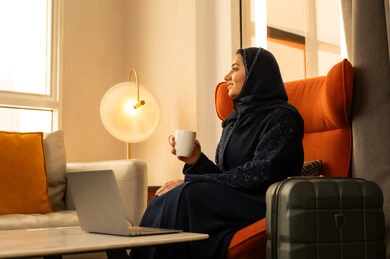 Enjoying warm winter herbs, expressions of pleasure and relaxation, spending leisure time sipping a favorite drink, an Arab Gulf Saudi woman wearing an abaya and hijab sitting in a hotel next to a suitcase drinking coffee, enjoying hot beverages.