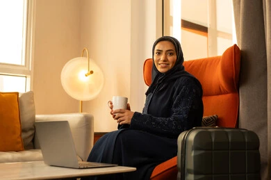 تعابير المتعة والاسترخاء، قضاء وقت الفراغ باحتساء المشروب المفضل, تناول الأعشاب الشتوية الدافئة، امراة عربية خليجية سعودية ترتدي العباءة والحجاب تجلس في الفندق بجانب حقيبة السفر تقوم بشرب القهوة، الاستمتاع بالمشروبات الساخنة