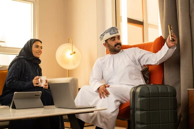 Documenting happy moments, gestures of joy and happiness, enjoying hot drinks, making a video call, integrating technology into daily routines, an Arab Gulf couple from Oman sitting inside a hotel next to a suitcase taking a selfie with a mobile phone.