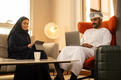 تعابير التركيز والسرور، تناول المشروبات الساخنة، العمل عن بعد، دمج التقنية والتكنولوجيا في الروتين اليومي، زوجان عربيان خليجيان عمانيان يجلسان داخل الفندق يعملان على الحاسب المحمول وجهاز التابلت