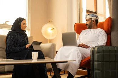 Drinking hot beverages, working remotely, integrating technology and tech into the daily routine, an Arab Gulf couple from Oman sitting inside a hotel working on a laptop and a tablet, expressions of focus and joy, exchanging chats and conversations.