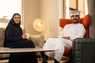 ايماءات السعادة و السرور، قضاء أوقات عائلية ممتعة، شرب القهوة العربية, التفاهم والترابط الفكري بين الزوجين، الجلوس في إحدى الفنادق الراقية، رجل عربي خليجي عماني يرتدي الدشداشة و الكمة يجلس بجانب حقيبة السفر برفقة زوجته