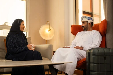 قضاء أوقات عائلية ممتعة، شرب القهوة العربية, التفاهم والترابط الفكري بين الزوجين، الجلوس في إحدى الفنادق الراقية، رجل عربي خليجي عماني يرتدي الدشداشة و الكمة يجلس بجانب حقيبة السفر يقوم بتبادل الأحاديث مع زوجته ، ايماءات السعادة و السرور