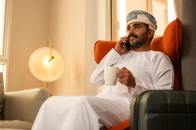 Making a phone call via mobile, chatting with family and friends, checking on relatives and hearing their news, enjoying a hot drink. An Arab Gulf Omani man wearing a dishdasha and kumma sits inside a hotel next to a suitcase holding a mobile phone and a cup of coffee.