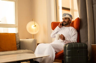 الدردشة مع الأهل والأصدقاء، اجراء مكالمة هاتفية عبر الجوال، الاطمئنان على الأقارب وسماع اخبارهم، الاستمتاع بالمشروب الساخن, رجل عربي خليجي عماني يرتدي الدشداشة و الكمة يجلس داخل الفندق بجانب حقيبة السفر يمسك بيده الهاتف المحمول و كوب من القهوه الدردشة مع الأهل والأصدقاء، اجراء مكالمة هاتفية عبر الجوال، الاطمئنان على الأقارب وسماع اخبارهم، الاستمتاع بالمشروب الساخن, رجل عربي خليجي عماني يرتدي الدشداشة و الكمة يجلس داخل الفندق بجانب حقيبة السفر يمسك بيده الهاتف المحمول و كوب من القهوه