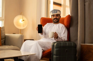 A black screen displayed on the mobile phone, enjoying a hot drink, using advanced modern technology, an Arab Gulf Omani man wearing a dishdasha and a kumma sitting inside the hotel next to a suitcase holding a mobile phone and a cup of coffee.