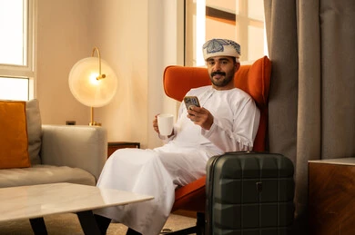 Enjoying a hot drink, using advanced modern technology, browsing social media on a mobile phone, an Arab Gulf Omani man wearing a dishdasha and kumma sitting inside a hotel next to a suitcase holding a mobile phone and a cup of coffee.